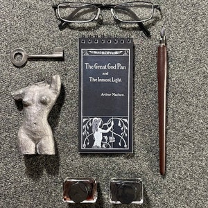Puede incluir: Un cuaderno negro y blanco con el título "The Great God Pan and The Inmost Light" de Arthur Machen. El cuaderno está rodeado de una llave de plata, una estatua de mujer de plata, unas gafas y una pluma de caligrafía marrón.