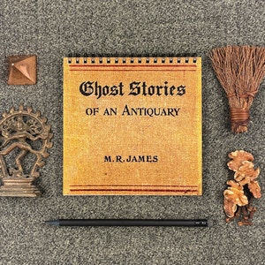 Peut inclure: Un livre vintage intitulé "Ghost Stories of an Antiquary" de M.R. James. Le livre a une couverture brune avec des lettres dorées et est relié avec une reliure à spirale.