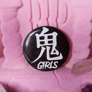 Op de afbeelding: Zwart-witte knop met een Japans karakter en het woord "GIRLS" in wit op een zwarte achtergrond.