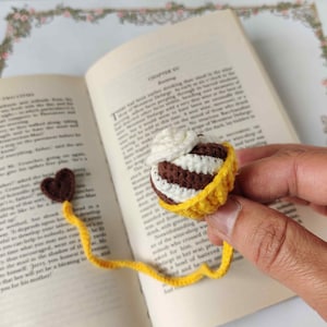 May include: A crocheted bookmark shaped like a chocolate and vanilla ice cream cone with a yellow crocheted base. The bookmark is attached to a yellow ribbon with a brown heart.