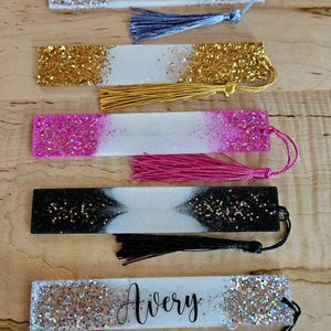 May include: A selection of handmade resin bookmarks with glitter and tassels. The bookmarks are in various colours, including silver, gold, pink, and black. One bookmark has the name "Avery" written on it.