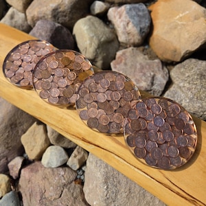 May include: Four circular coasters made of clear resin embedded with numerous copper-coloured pennies. Each coaster features a smooth, glossy finish with the metallic coins arranged in a dense, overlapping pattern throughout the transparent material.