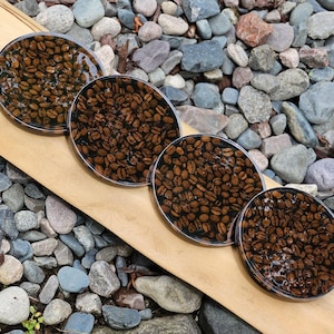 Peut inclure: Quatre dessous de verre ronds avec une finition en résine transparente, chacun rempli de grains de café. Les dessous de verre sont disposés sur une surface en bois, entourés de petits cailloux.