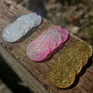May include: Three sets of glittery car coasters in silver, pink, and gold. The coasters are round with a small notch for easy removal. The coasters are arranged on a weathered wooden surface.
