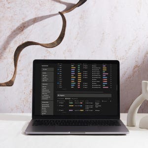 May include: A laptop displaying a dark-themed project management interface. The screen shows a list of tasks with color-coded progress bars. The laptop is open on a white surface, with a decorative element and a marble-patterned background.