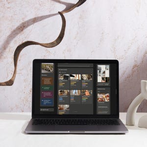 May include: A silver laptop displaying a digital interface with various icons and text. The screen shows a dark-themed application with multiple image thumbnails and text. A white ceramic decorative object is on the right. The background features a marble wall and a curved brown branch.