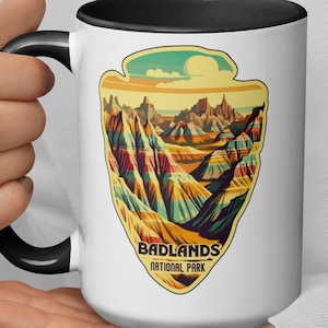 May include: White ceramic mug with a black rim and handle. The mug features a colorful illustration of Badlands National Park with the text "Badlands National Park".