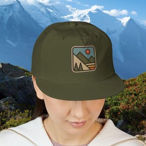 Puede incluir: Gorra de b&eacute;isbol verde oliva con un parche cuadrado con un dise&ntilde;o de paisaje monta&ntilde;oso con un sol y formas geom&eacute;tricas. La gorra tiene una visera plana.