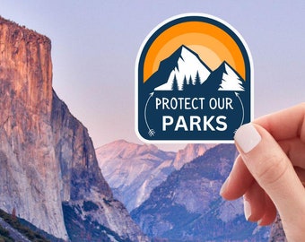 May include: A sticker with the text "PROTECT OUR PARKS" and a mountain range design, held in front of a scenic mountain landscape. The sticker has a blue and white border with a yellow and orange sun and mountain graphic.