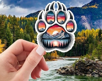 Puede incluir: Una pegatina de huella de pata de oso blanca con un paisaje de puesta de sol colorido en su interior. El paisaje presenta montañas, un lago y árboles. La pegatina se sostiene en una mano contra un fondo de río y árboles.