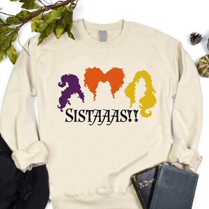 May include: Cream-colored sweatshirt with the word "Sistaaas!!" and three stylized witch hair silhouettes in purple, orange, and yellow. The sweatshirt is surrounded by Halloween decorations, including pumpkins and a skull.