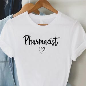 May include: White t-shirt with the word "Pharmacist" in black script and a small heart below. The shirt is hanging on a wooden hanger with other clothing items in the background.