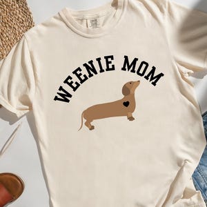 May include: Cream-colored t-shirt with the text "WEENIE MOM" above a brown dachshund illustration with a black heart. The shirt is paired with denim shorts, sandals, and a straw hat.