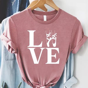 May include: A dusty rose t-shirt with the word "LOVE" in large white letters. Two white ballet slippers are above the letter "E". The shirt is hanging on a wooden hanger with other clothing.