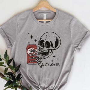 Op de afbeelding: Een grijs gem&ecirc;leerd T-shirt met korte mouwen en een zwart-witte afbeelding van een menselijke schedel die een rood frisdrankblikje met het label Pepper vasthoudt. Het ontwerp bevat kleine zwarte sterren en de cursieve tekst 'til death. Het shirt hangt aan een hanger.