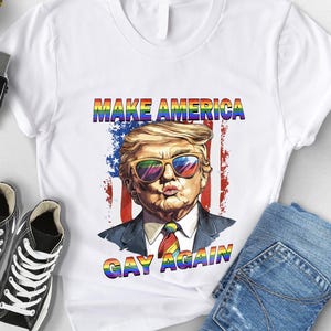 May include: White t-shirt featuring a graphic of a man wearing rainbow sunglasses, with the text "MAKE AMERICA GAY AGAIN" in rainbow colors. The shirt is paired with blue jeans and black sneakers.