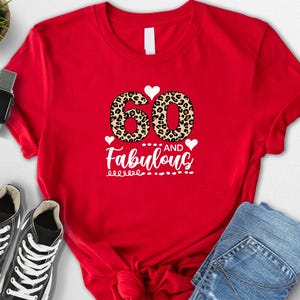 May include: A red t-shirt with the text "60 and Fabulous" in white script and leopard print numbers. The shirt is styled with black sneakers, a smartwatch, and blue jeans.