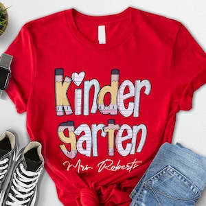 K&ouml;nnte beinhalten: Ein rotes T-Shirt mit dem Wort "Kindergarten" in einer farbenfrohen, verspielten Schriftart, mit einem Herz &uuml;ber dem "i". Das Shirt hat auch den Namen "Mrs. Roberts" unter dem Wort "garten" aufgedruckt. Schwarze Turnschuhe und blaue Jeans sind ebenfalls im Bild.