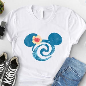 May include: White t-shirt featuring a blue Mickey Mouse head design with a flower and wave pattern. The shirt is paired with black sneakers, blue jeans, and a smartwatch. A small plant and cotton branch are also visible.