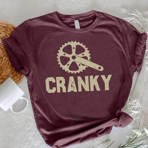 May include: A maroon short-sleeved t-shirt featuring a cream-colored graphic of a bicycle crankset and chainring centered on the chest. Below the graphic, the word CRANKY is printed in a bold, distressed, cream-colored sans-serif font.