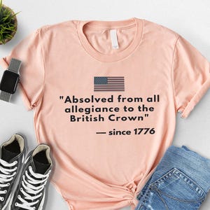 May include: A peach-colored t-shirt with the American flag and the text "Absolved from all allegiance to the British Crown - since 1776." The shirt is paired with blue jeans and black sneakers.