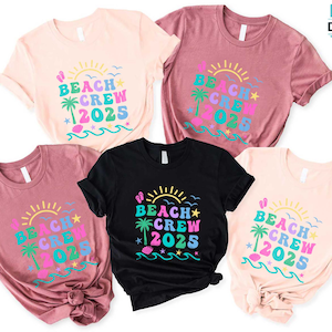 Puede incluir: Cinco camisetas de manga corta en varios colores: rosa claro, malva y negro. Cada camiseta presenta el texto "BEACH CREW 2025" en un dise&ntilde;o colorido con un sol, una palmera y olas. Las camisetas est&aacute;n bien dispuestas.