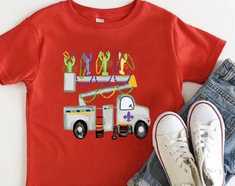 Puede incluir: Una camiseta roja con un diseño de camión que transporta cangrejos de río y cuentas de Mardi Gras. El camión tiene una flor de lis morada. La camiseta se combina con pantalones cortos de mezclilla y zapatillas blancas.