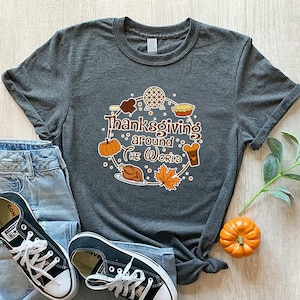 May include: A gray t-shirt with the text "Thanksgiving Around The World" in a circular design with pumpkins, a turkey, and other fall-themed graphics. The shirt is paired with blue jeans and black sneakers.