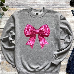 May include: A gray sweatshirt featuring a large, sparkly pink bow graphic. The bow has a textured appearance and is centered on the front of the long-sleeved top. Black and white sneakers and blue jeans are also in the image.
