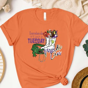 May include: Orange t-shirt with a Mardi Gras theme. The design features a white boot filled with festive items, a green alligator, and the text "Everywhere else it's just a Tuesday Mardi Gras."