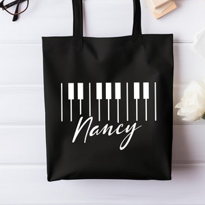 May include: Black tote bag with a white piano keyboard design and the name "Nancy" printed below.