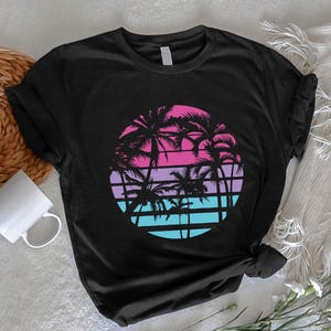 May include: Black t-shirt featuring a circular graphic of palm trees against a gradient sunset in pink, purple, and blue. The design includes horizontal lines and silhouettes of palm trees, evoking a tropical theme.