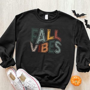 May include: Black sweatshirt with the text "FALL VIBES" in a colorful, retro font. The sweatshirt is paired with white sneakers with red accents. A small pumpkin and paper bats are also in the image.