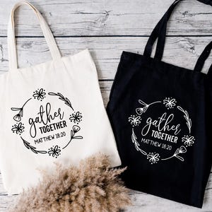 May include: Two tote bags, one cream and one black, with the words "gather together" and "Matthew 18:20" printed in a floral wreath design. The bags have long handles and are made of a sturdy fabric.