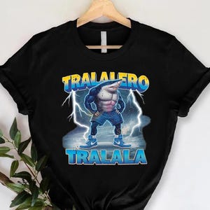 May include: Black t-shirt featuring a muscular shark character with the text "TRALALERO TRALALA" in blue and yellow. The shark is depicted with a human-like physique, wearing blue jeans and sneakers, with lightning bolts in the background.