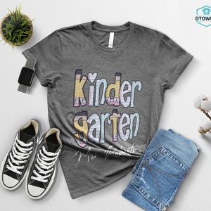 K&ouml;nnte beinhalten: Ein meliertes graues T-Shirt mit dem Wort "Kindergarten" in Pastellfarben und Bleistiftakzenten. Das Bild zeigt blaue Jeans, schwarze Turnschuhe und eine Smartwatch. Eine kleine Pflanze und ein Baumwollzweig sind ebenfalls zu sehen.