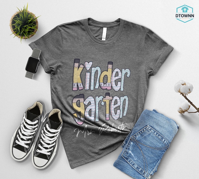 K&ouml;nnte beinhalten: Ein meliertes graues T-Shirt mit dem Wort "Kindergarten" in Pastellfarben und Bleistiftakzenten. Das Bild zeigt blaue Jeans, schwarze Turnschuhe und eine Smartwatch. Eine kleine Pflanze und ein Baumwollzweig sind ebenfalls zu sehen.