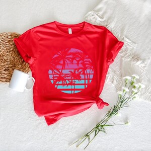May include: A red t-shirt featuring a circular graphic of palm trees against a sunset backdrop. The design incorporates shades of pink, blue, and purple. The shirt is made of a soft material and has short sleeves.