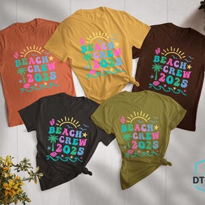 Puede incluir: Cinco camisetas de colores variados, naranja, amarillo, marr&oacute;n, negro y verde oliva, con el texto "BEACH CREW 2025". El dise&ntilde;o incluye un sol, una palmera y olas, con tem&aacute;tica playera.