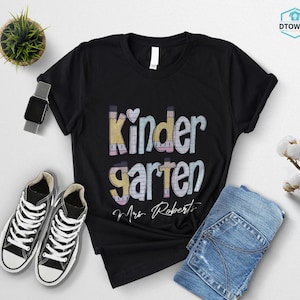 K&ouml;nnte beinhalten: Schwarzes T-Shirt mit dem Wort "Kindergarten" in einem farbenfrohen Design mit Bleistiften und dem Schriftzug "Mrs. Roberts". Das Shirt wird mit blauen Jeans und schwarz-wei&szlig;en Turnschuhen kombiniert.