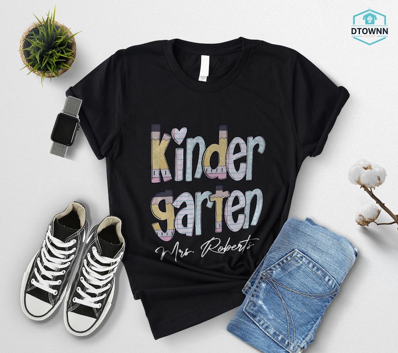 K&ouml;nnte beinhalten: Schwarzes T-Shirt mit dem Wort "Kindergarten" in einem farbenfrohen Design mit Bleistiften und dem Schriftzug "Mrs. Roberts". Das Shirt wird mit blauen Jeans und schwarz-wei&szlig;en Turnschuhen kombiniert.