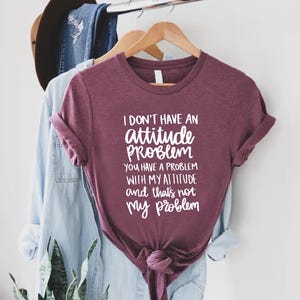 K&ouml;nnte beinhalten: Burgunderfarbenes T-Shirt mit dem wei&szlig;en Text: "I don't have an attitude problem, you have a problem with my attitude and that's not my problem". Das T-Shirt h&auml;ngt an einem Holzkleiderb&uuml;gel.