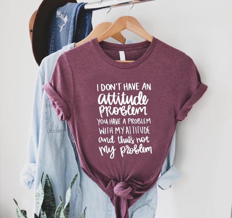 K&ouml;nnte beinhalten: Burgunderfarbenes T-Shirt mit dem wei&szlig;en Text: "I don't have an attitude problem, you have a problem with my attitude and that's not my problem". Das T-Shirt h&auml;ngt an einem Holzkleiderb&uuml;gel.