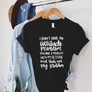 K&ouml;nnte beinhalten: Schwarzes T-Shirt mit wei&szlig;em Text: "I DON'T HAVE AN attitude PROBLEM YOU HAVE A PROBLEM WITH MY ATTITUDE and that's not my problem." Das Shirt h&auml;ngt an einem Holzkleiderb&uuml;gel.