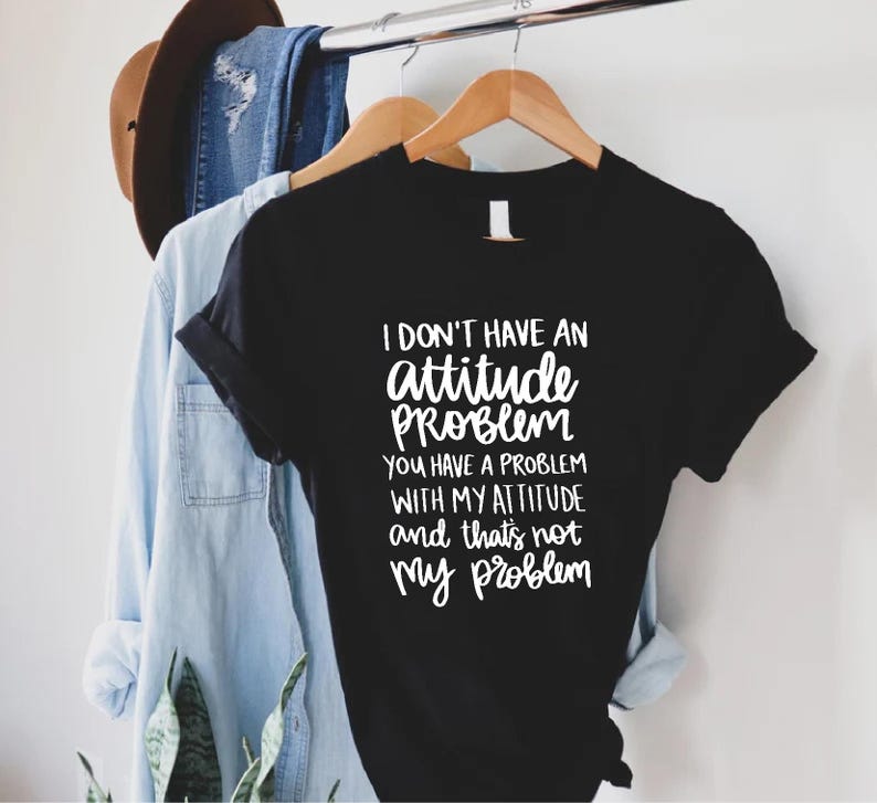 K&ouml;nnte beinhalten: Schwarzes T-Shirt mit wei&szlig;em Text: "I DON'T HAVE AN attitude PROBLEM YOU HAVE A PROBLEM WITH MY ATTITUDE and that's not my problem." Das Shirt h&auml;ngt an einem Holzkleiderb&uuml;gel.