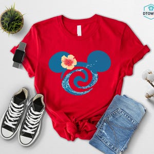 May include: A red t-shirt featuring a blue Mickey Mouse head design with a hibiscus flower and wave pattern. The shirt is paired with black sneakers, a smartwatch, and blue jeans. A small potted plant is also visible.