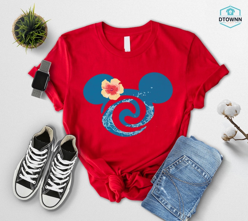 May include: A red t-shirt featuring a blue Mickey Mouse head design with a hibiscus flower and wave pattern. The shirt is paired with black sneakers, a smartwatch, and blue jeans. A small potted plant is also visible.