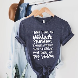 K&ouml;nnte beinhalten: Ein marineblaues T-Shirt mit dem Text "I DON'T HAVE AN attitude PROBLEM YOU HAVE A PROBLEM WITH MY ATTITUDE and that's not my problem" in Wei&szlig;. Das Shirt h&auml;ngt an einem Holzb&uuml;gel mit anderer Kleidung.
