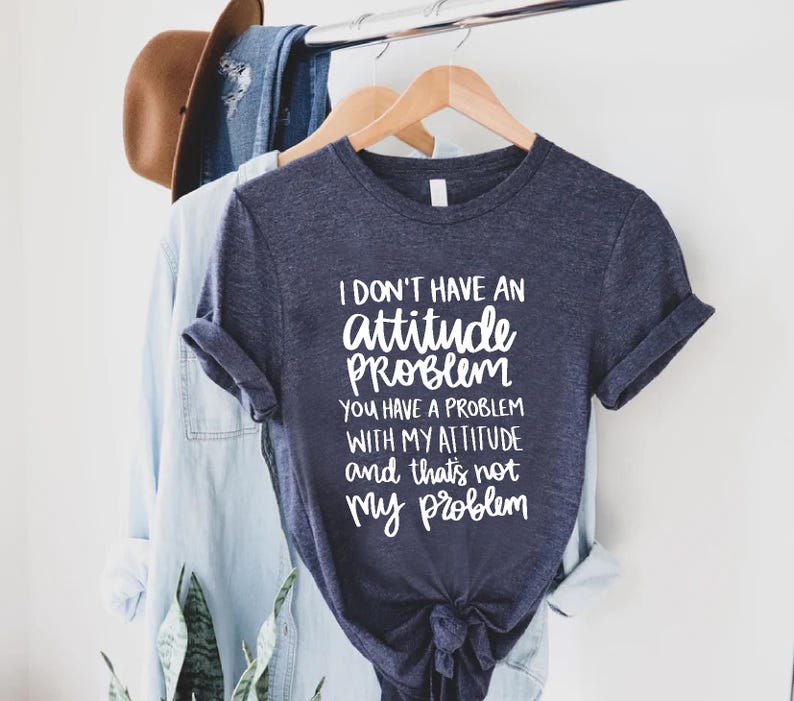 K&ouml;nnte beinhalten: Ein marineblaues T-Shirt mit dem Text "I DON'T HAVE AN attitude PROBLEM YOU HAVE A PROBLEM WITH MY ATTITUDE and that's not my problem" in Wei&szlig;. Das Shirt h&auml;ngt an einem Holzb&uuml;gel mit anderer Kleidung.