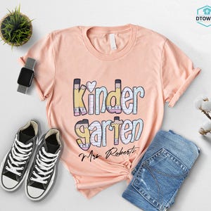 K&ouml;nnte beinhalten: Pfirsichfarbenes T-Shirt mit dem Wort "Kindergarten" in einem farbenfrohen Design im Bleistift-Stil. Der Name "Mrs. Roberts" ist darunter aufgedruckt. Das Shirt wird mit blauen Jeans und schwarzen Turnschuhen kombiniert.
